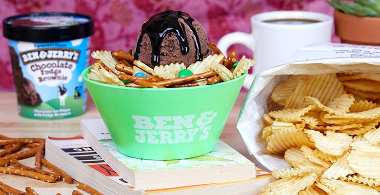 A pint of ice cream, pretzels, potato chips and candy on desk along with the finished sundae